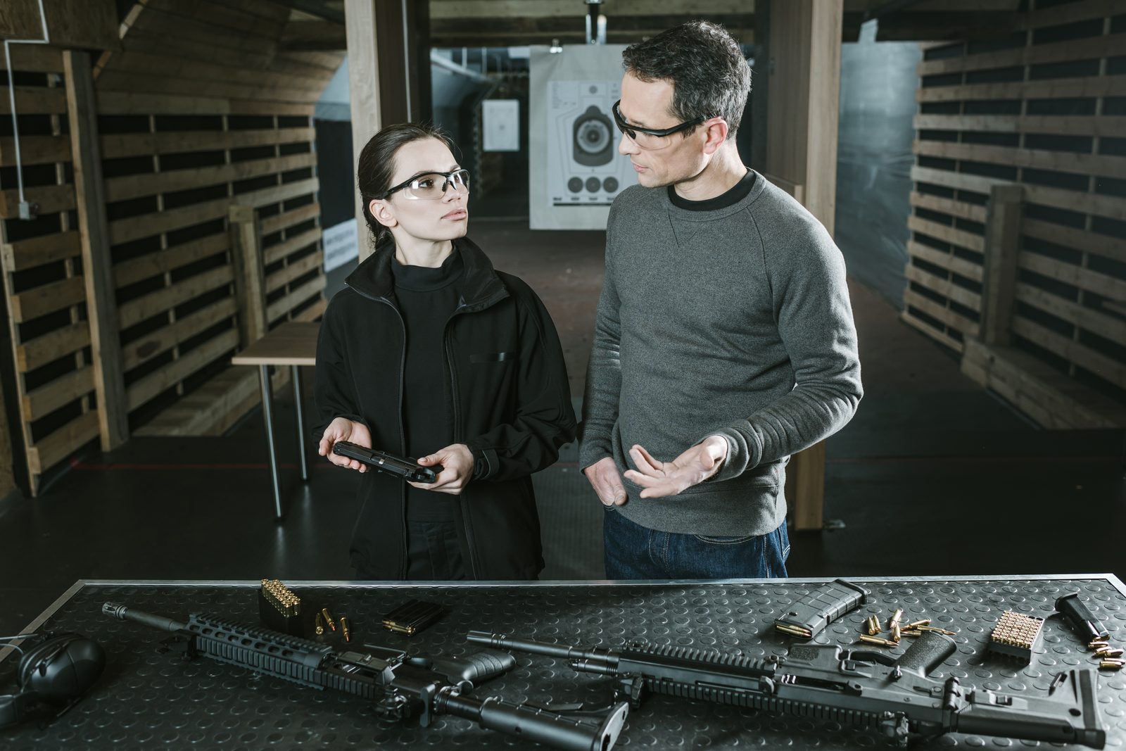 Shooting instructor talking with a student at an indoor range, with rifles and ammunition on the bench